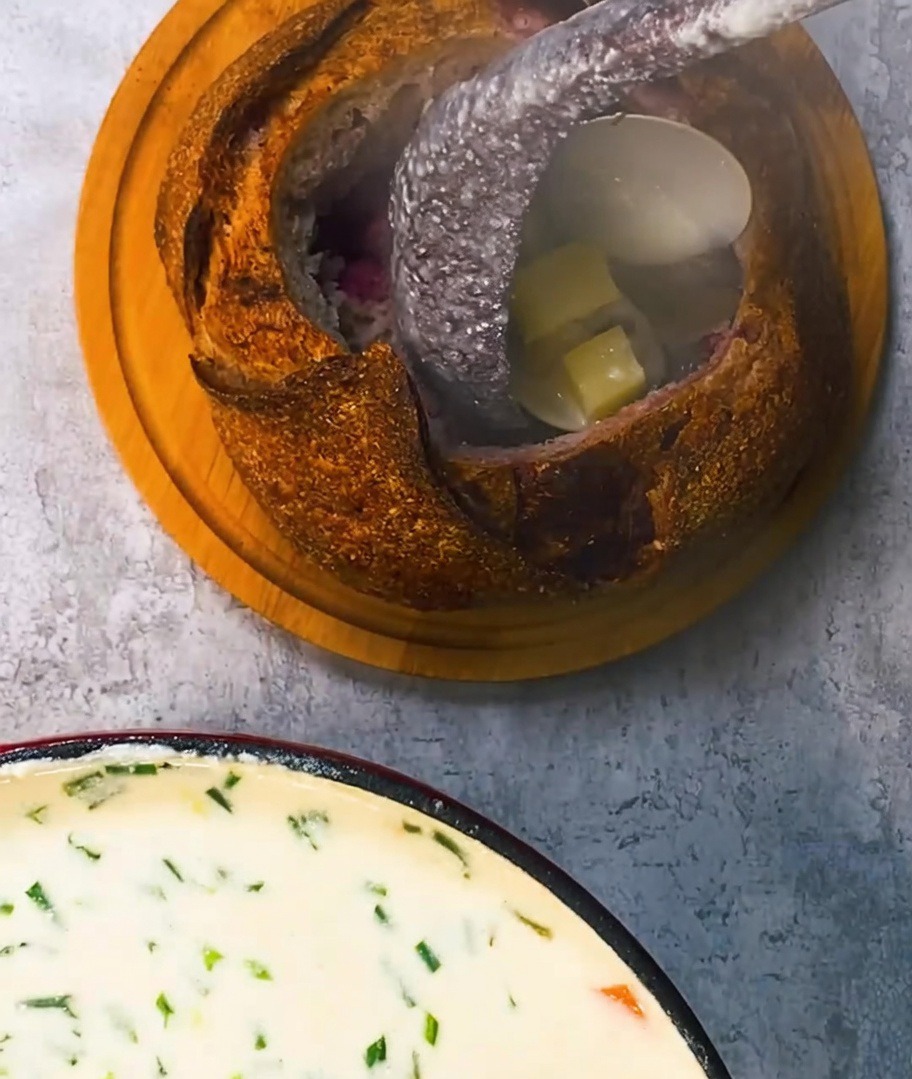 Sopa de almejas cremosa siendo servida en el cuenco de pan y decorada con tocino crujiente.
