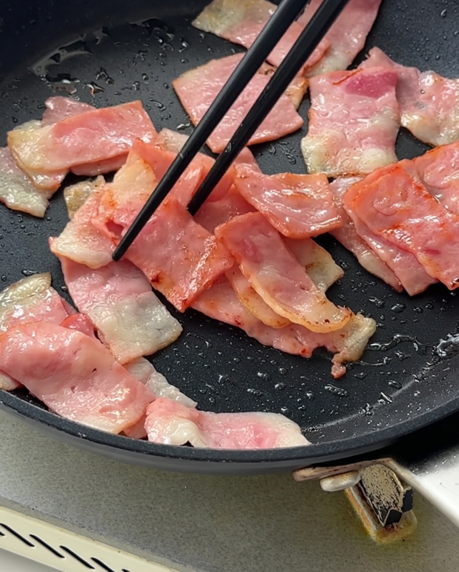 Cebollas moradas picadas siendo integradas en la sartén con el tocino dorado.