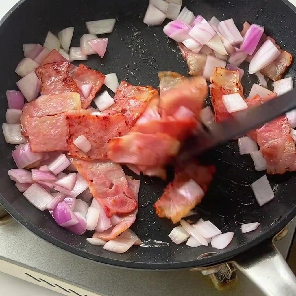 Pasta fusilli y tallos de espárragos verde brillante siendo incorporados en una sartén con tocino dorado y cebollas picadas.