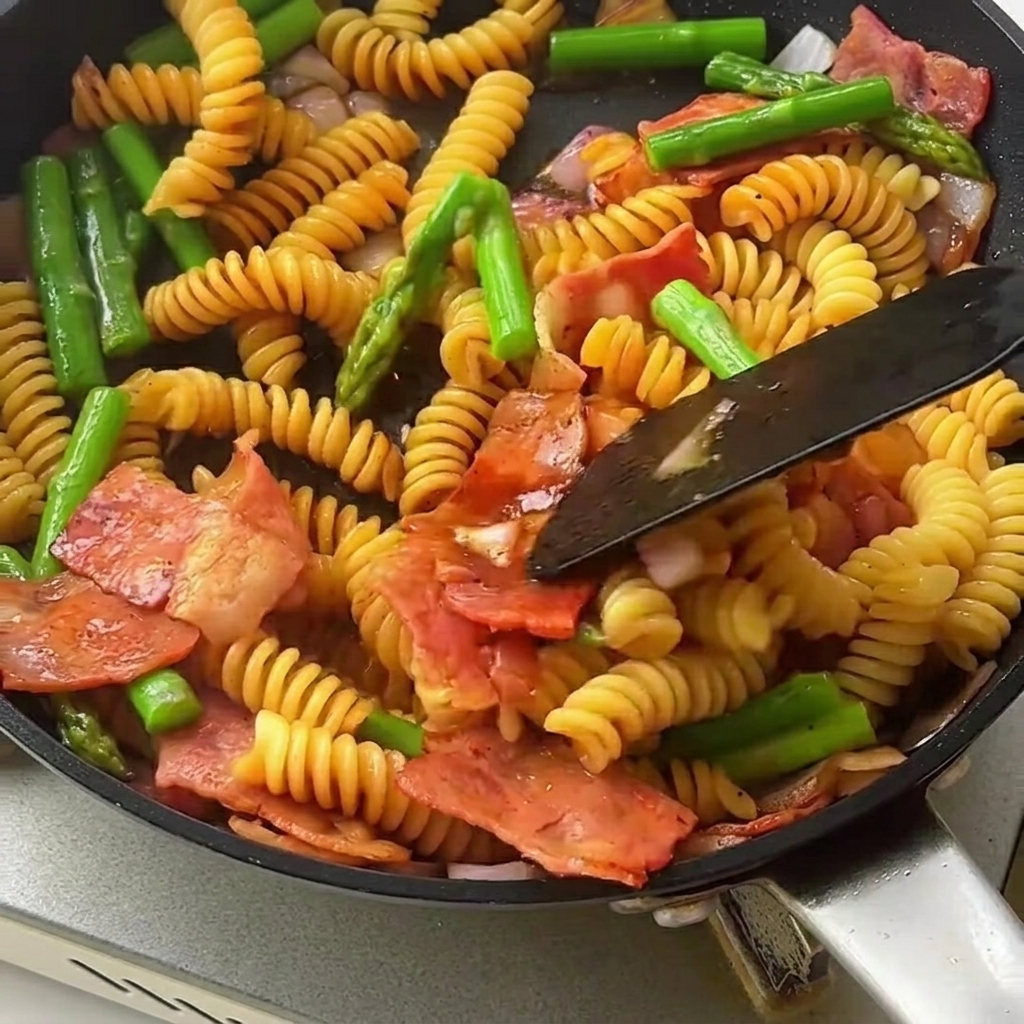 Una espátula mezclando la pasta fusilli glaseada con salsa, espárragos crujientes y trozos de tocino en una sartén grande.
