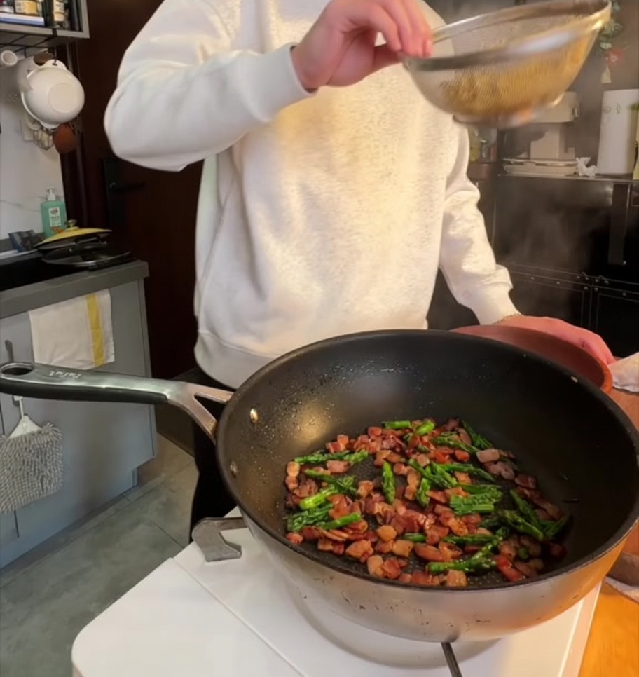 Pasta penne cocida siendo añadida a una sartén con panceta crujiente y puntas de espárrago salteadas.