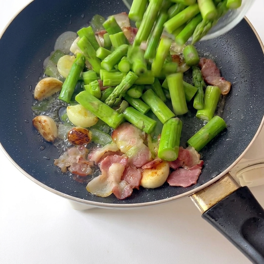 Trozos de espárragos blanqueados vertidos en una sartén con ajo frito y bacon dorado.