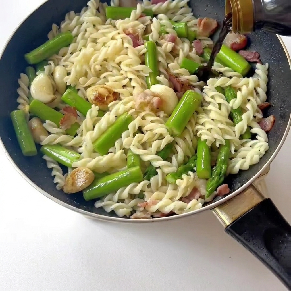 Primer plano de una espátula removiendo pasta, bacon y espárragos mientras se añade salsa de soja ligera.