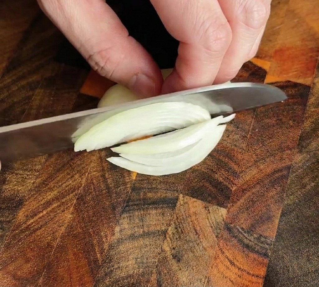 Cuchillo de chef cortando una cebolla en tiras finas sobre una tabla de madera.