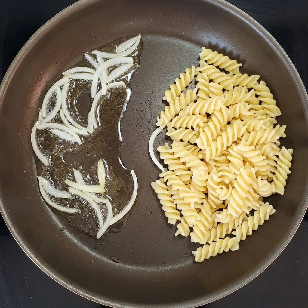 Pasta en espiral siendo mezclada con cebollas salteadas en una sartén.