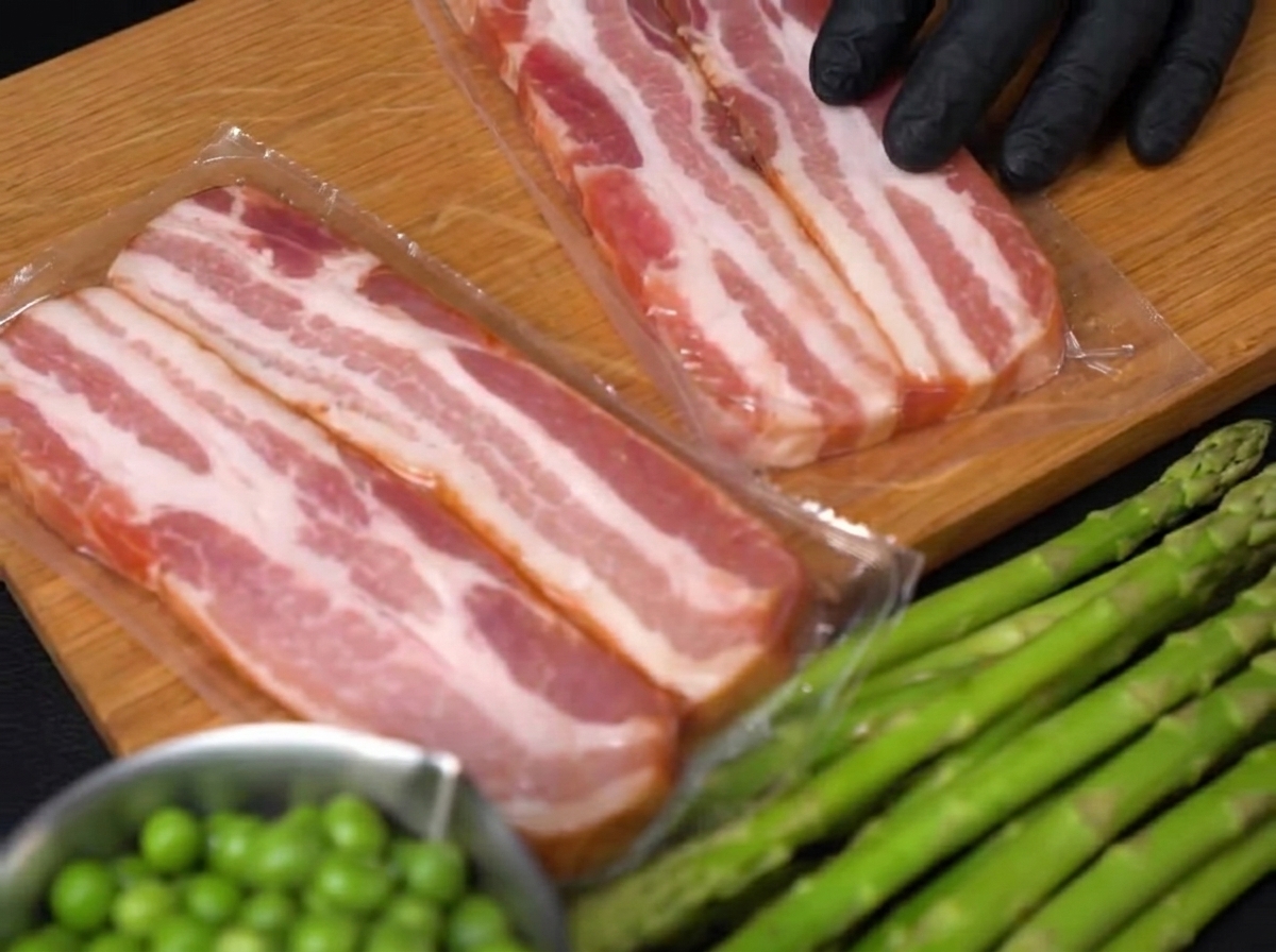 Primer plano de una mano enguantada usando un cuchillo para cortar tiras gruesas de tocino crudo sobre una tabla de madera.