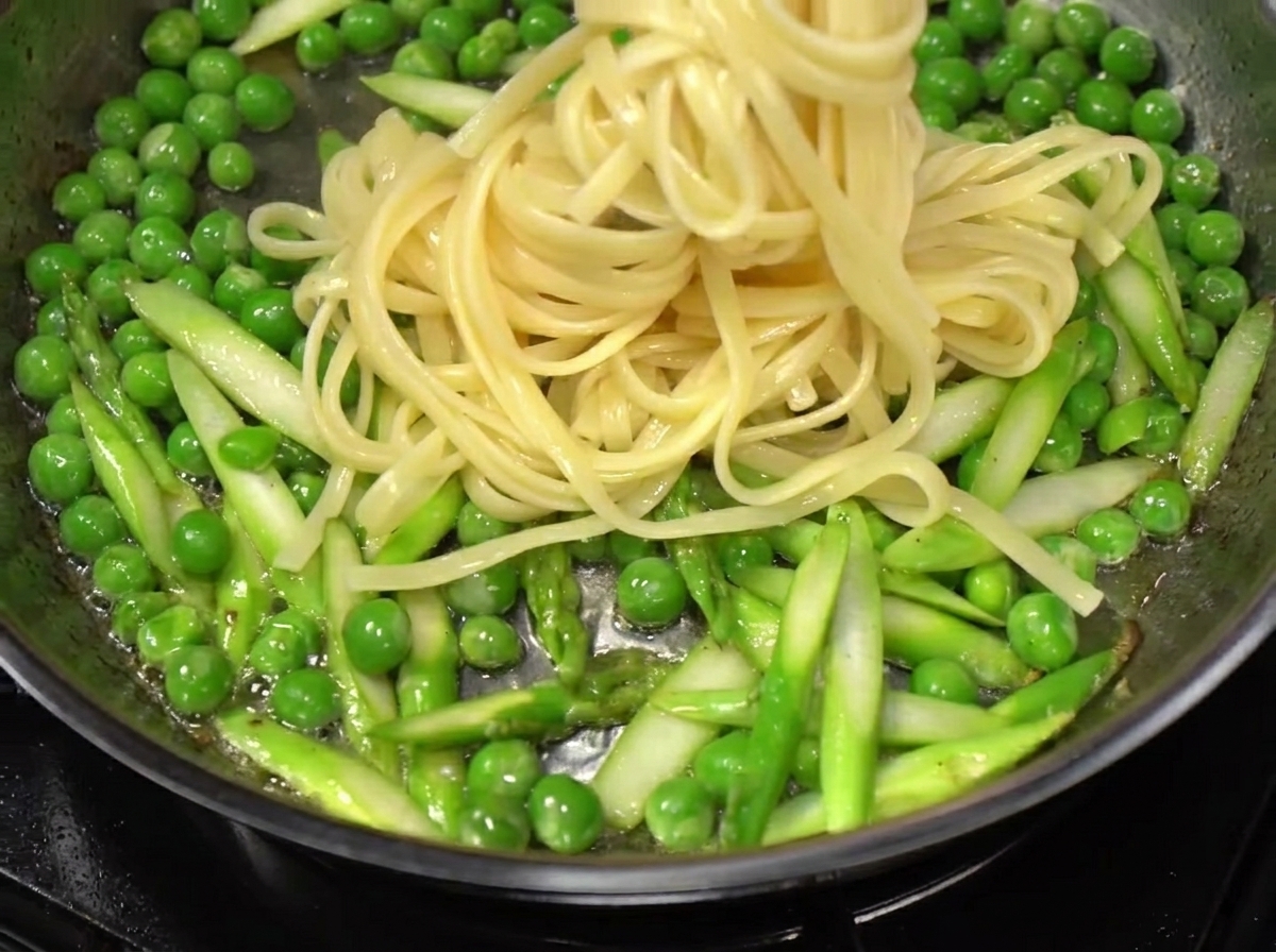 Espaguetis siendo retirados del agua hirviendo y añadidos a una sartén con verduras salteadas y tocino.