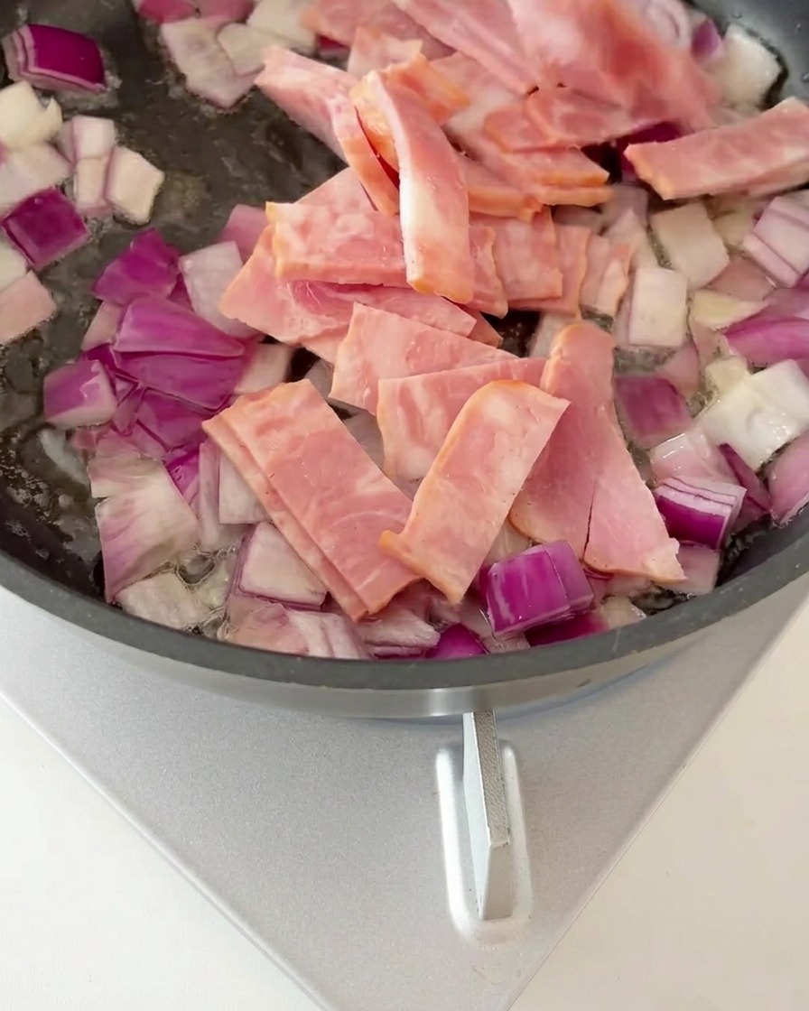 Trozos de bacon mezclados con cebolla sofrita en una sartén, empezando a dorarse.