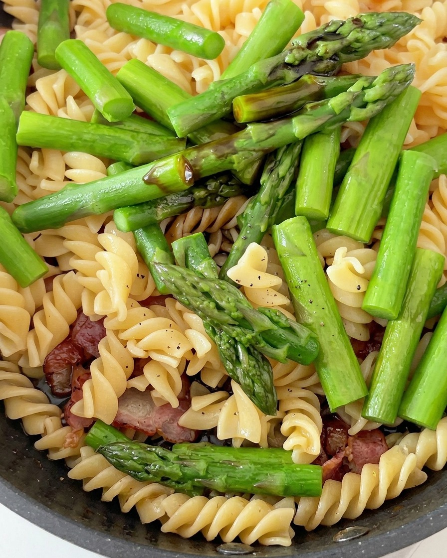 Sartén que contiene pasta fusilli cocida, espárragos verdes vibrantes y bacon cocinado, siendo condimentados con salsas.
