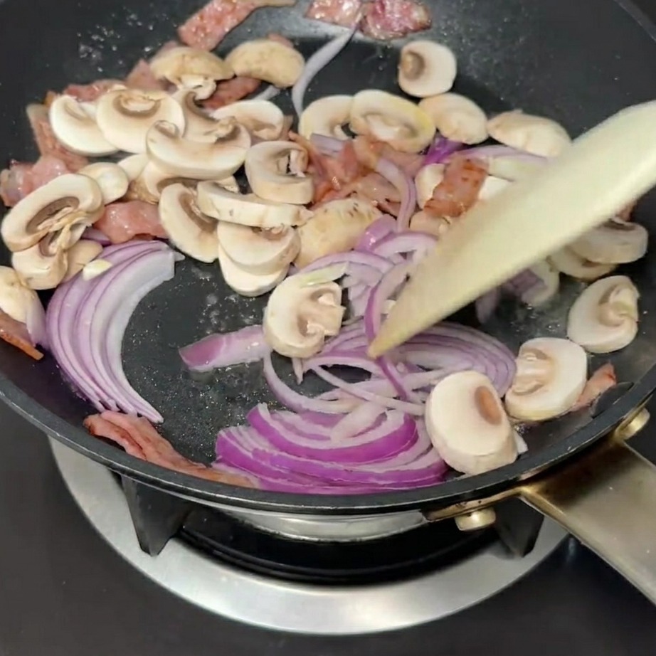 Cebolla roja en rodajas y champiñones siendo salteados en una sartén con bacon.
