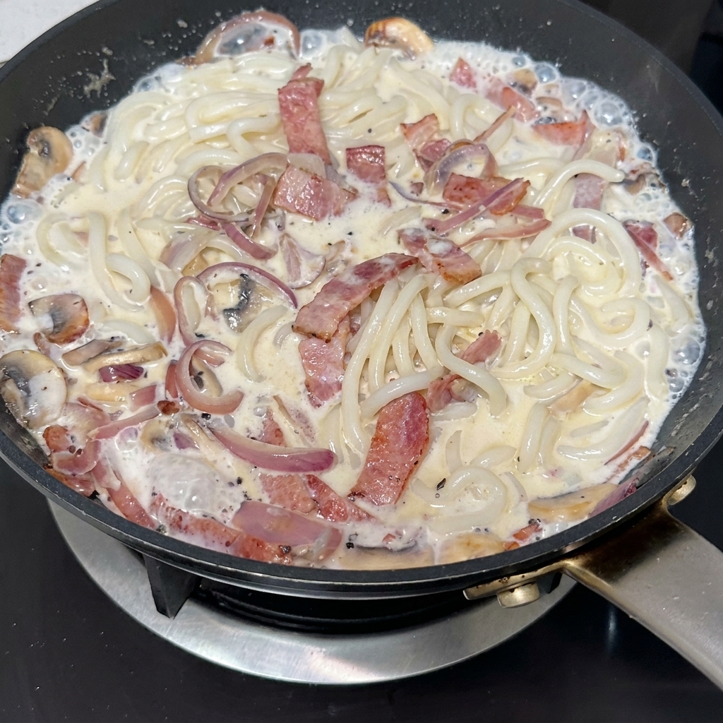 Pasta de trufa negra siendo incorporada a una sartén con bacon, champiñones y cebolla caramelizada.