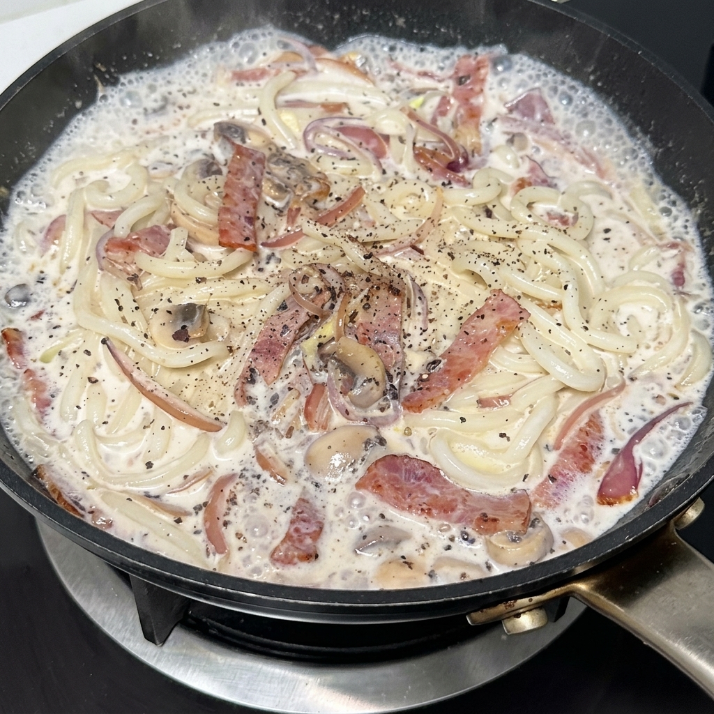 Una sartén de udon cremoso con bacon y champiñones siendo sazonada con pimienta negra.