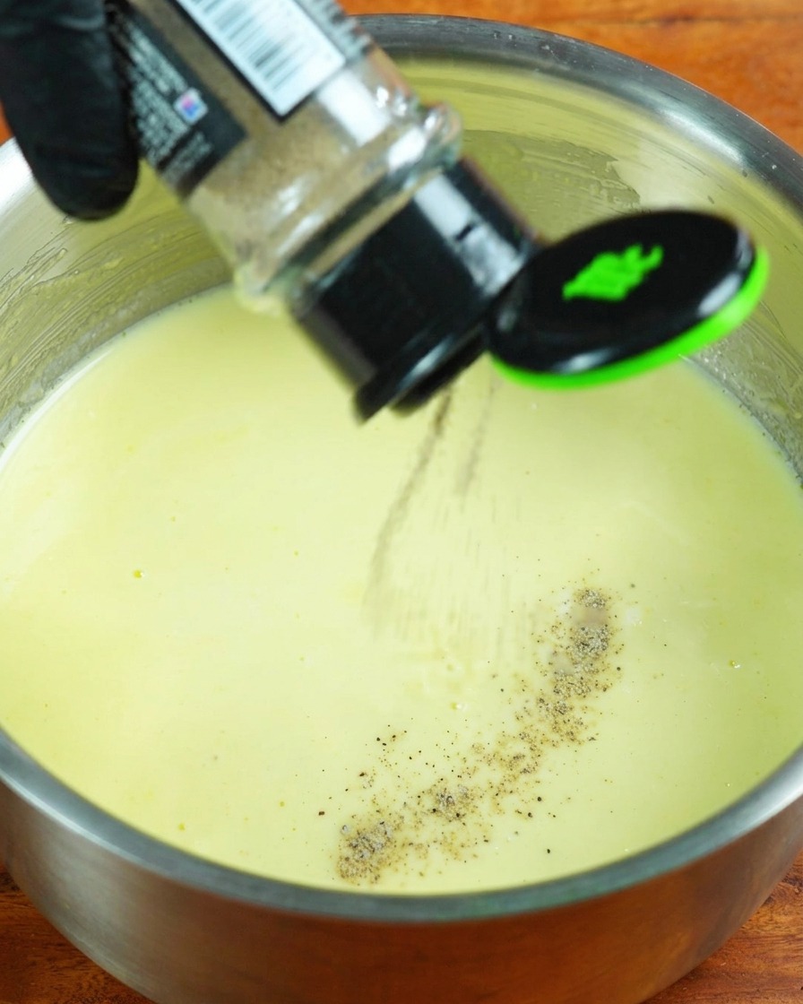 Nata, pimienta blanca y sal siendo añadidas a la salsa de leche a fuego lento.