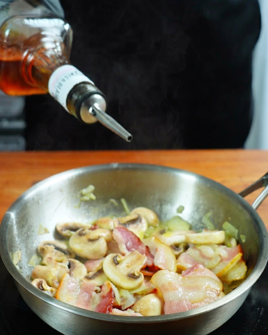 Whisky siendo vertido de una botella a una sartén con bacon y champiñones.