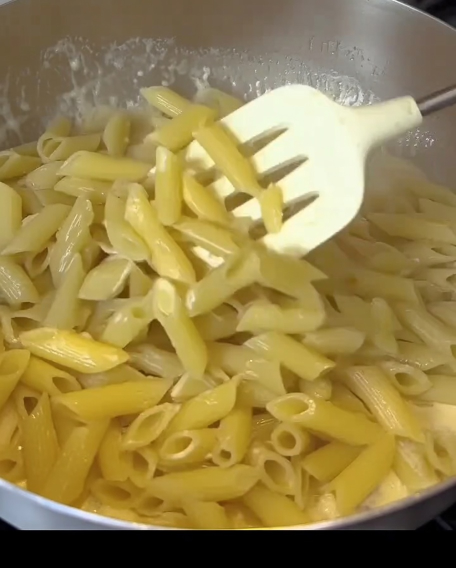 Pasta penne siendo mezclada en una salsa blanca y cremosa con trozos de tocino en una sartén.