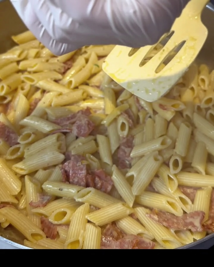 Manos con guantes espolvoreando sazonador sobre una sartén de pasta cremosa con tocino.