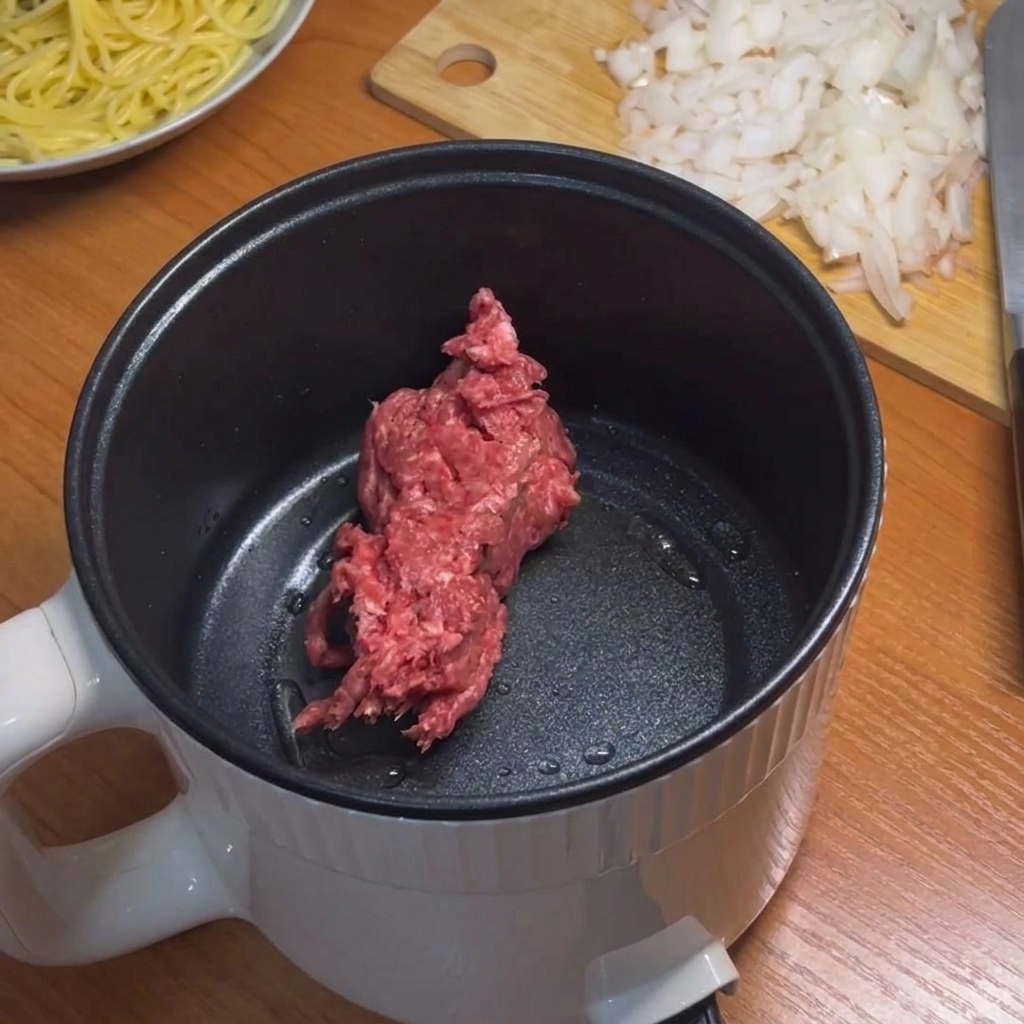 Carne picada siendo dorada en una pequeña olla eléctrica con una espátula de silicona azul claro.