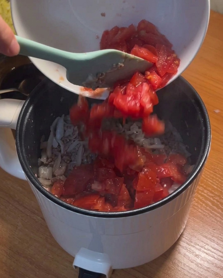 Tomates picados siendo vertidos desde un cuenco blanco a la olla con la carne y la cebolla sofritas.