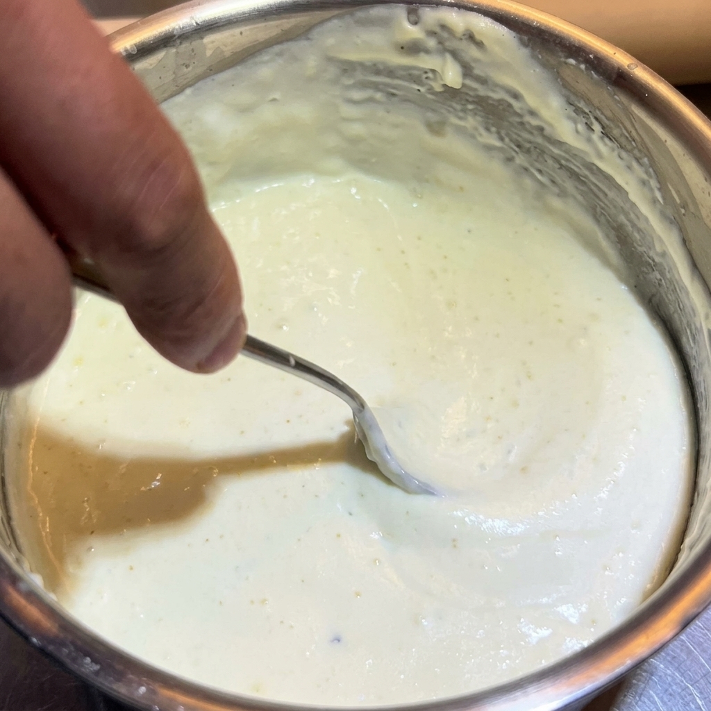 Una cuchara de metal revolviendo una salsa blanca de queso espesa y cremosa en una olla de acero inoxidable.
