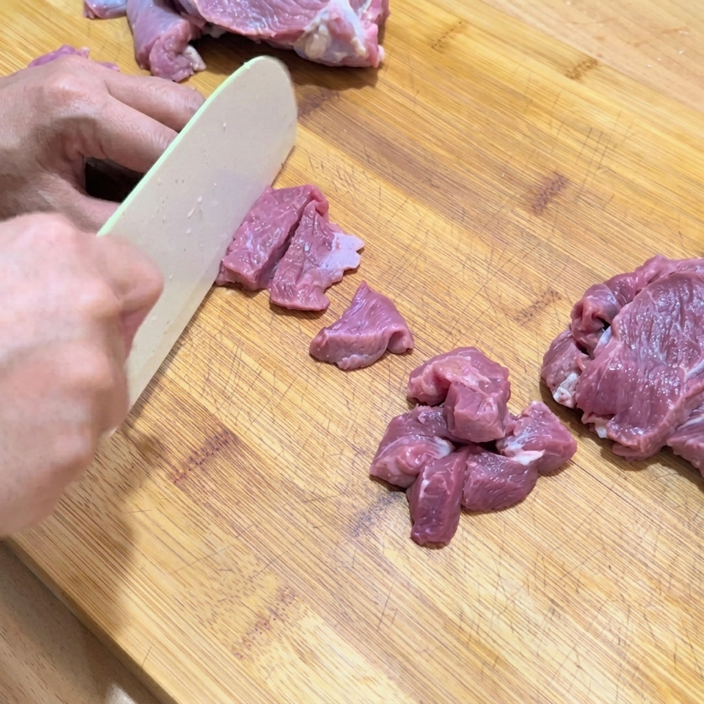 Manos cortando ternera cruda en cubos pequeños y uniformes sobre una tabla de cortar.