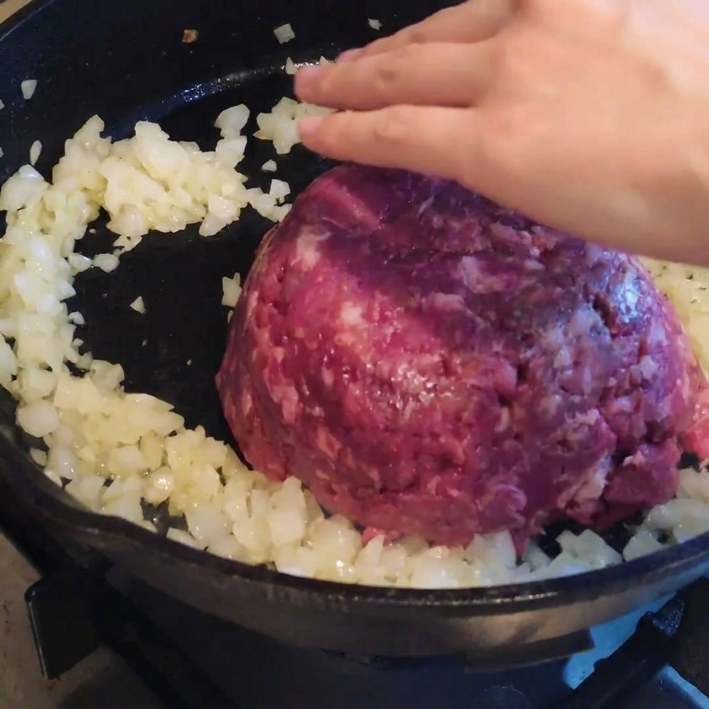 Carne picada introduciéndose en una sartén con las cebollas previamente sofritas, lista para remover y sazonar.