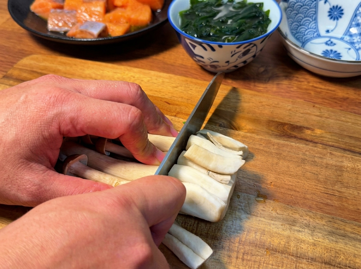 Vista cercana de un cuchillo cortando las raíces de un manojo de setas shimeji sobre una tabla de cortar de madera.