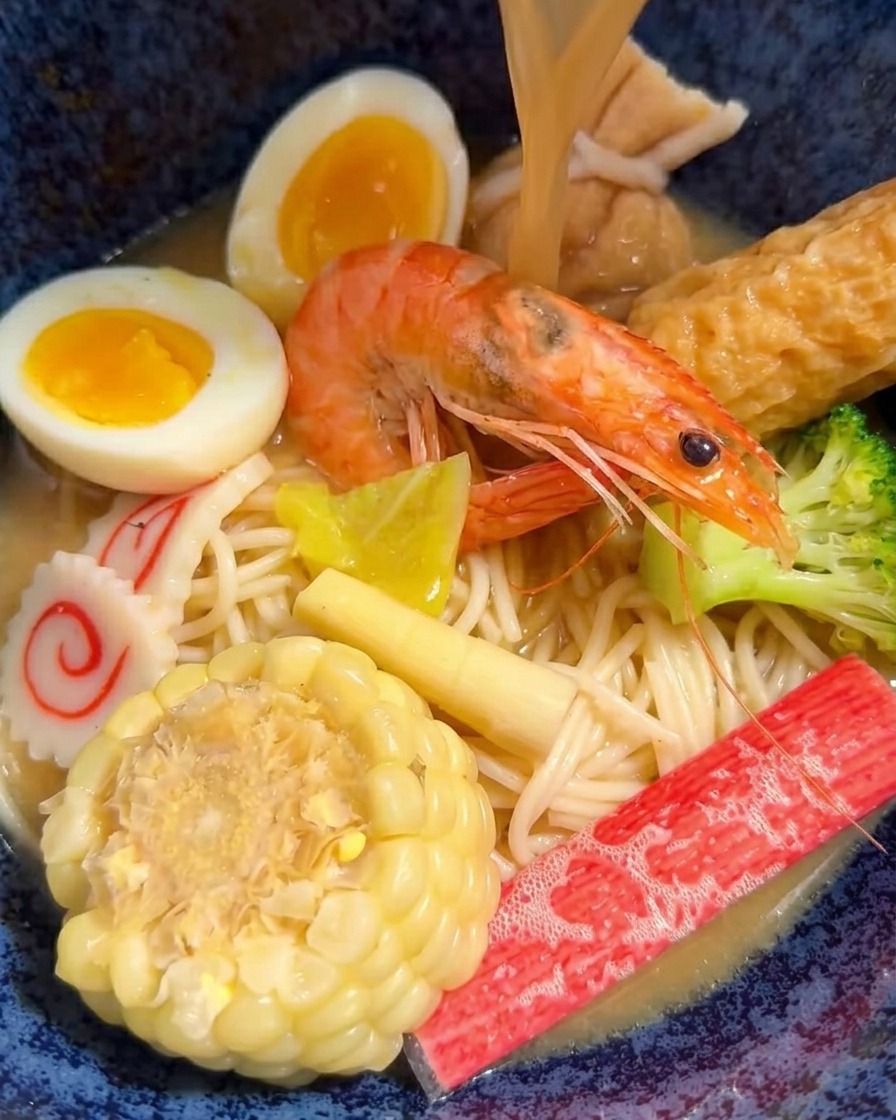 Caldo de miso caliente siendo vertido con un cucharón en un tazón de ramen lleno de carnes y vegetales.
