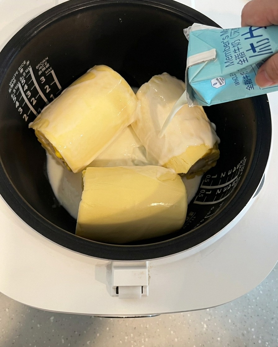 Leche vertiéndose desde un cartón en la olla de una arrocera que contiene maíz y queso.
