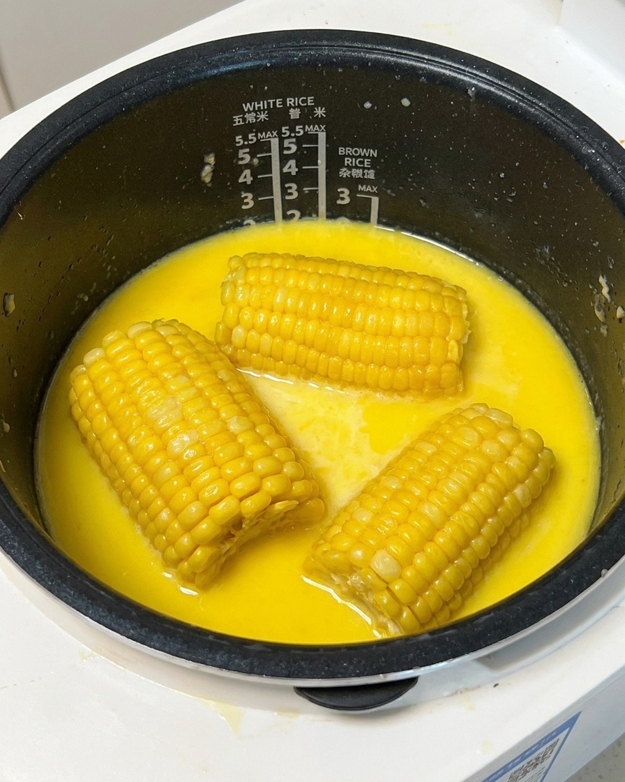 Un trozo de maíz amarillo cocido siendo levantado de un caldo lechoso en una arrocera usando un pincho de madera.