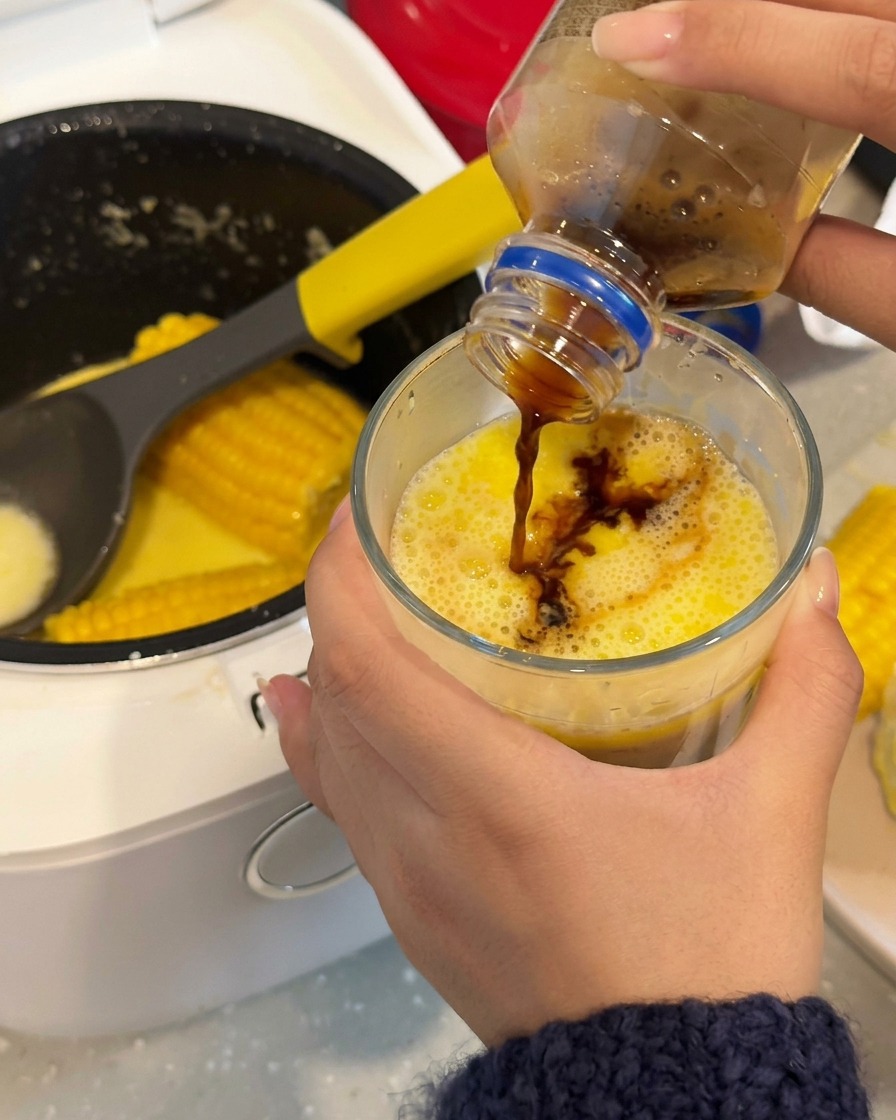 Café oscuro vertiéndose en un vaso que contiene un caldo de maíz lechoso de color amarillo brillante.