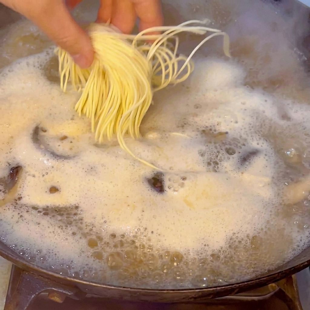 Una mano dejando caer con cuidado un manojo de fideos amarillos y frescos de ramen en el caldo de mariscos hirviendo.