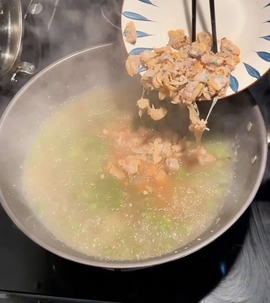 Carne de almeja siendo añadida de un plato a una olla de caldo hirviendo sazonado con vegetales verdes.