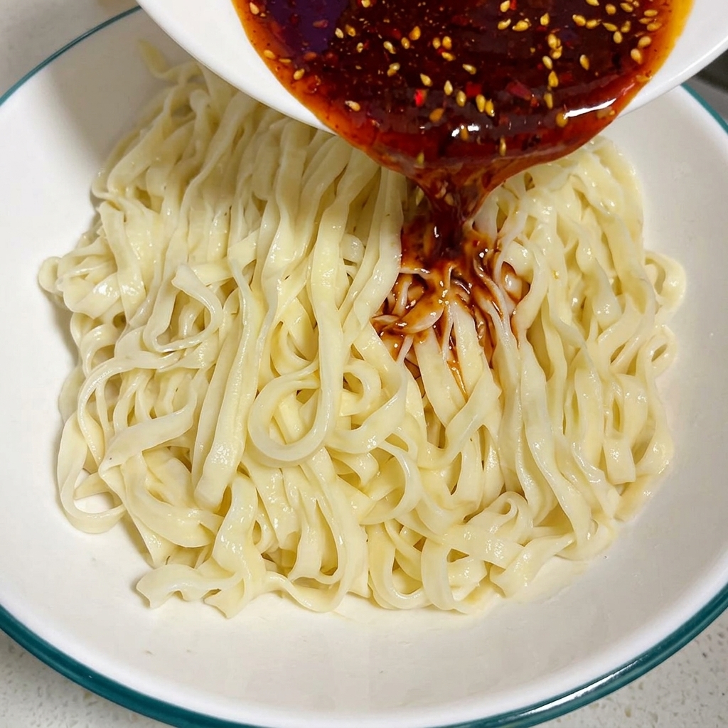 Una rica salsa roja oscura de chile y curry vertiéndose desde un tazón pequeño sobre una cama de fideos planos cocidos en un tazón más grande.