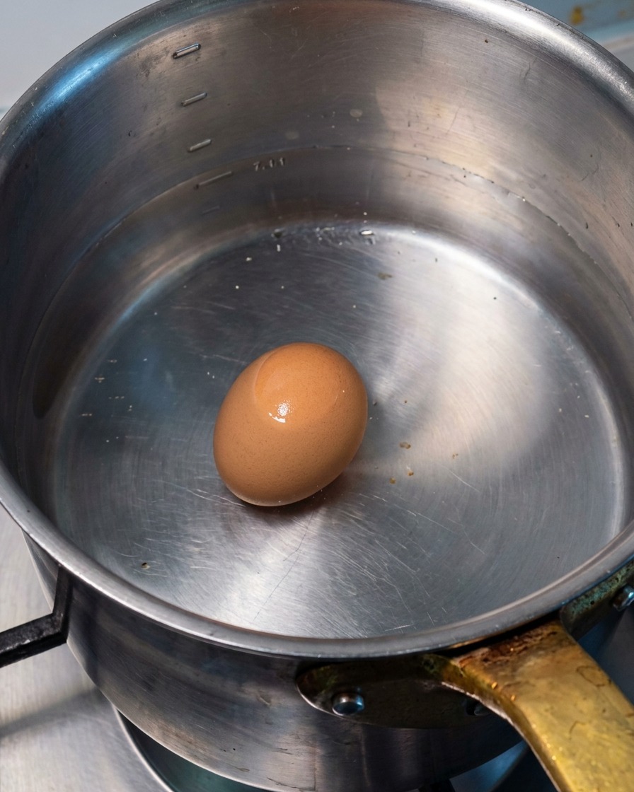 Un huevo entero reposando en el fondo de una olla de metal llena de agua caliente.