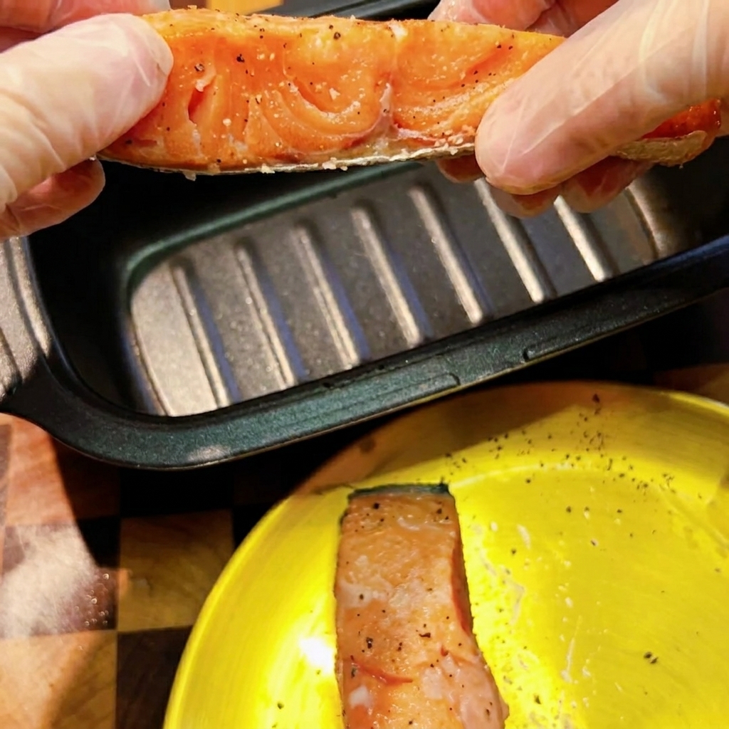 Manos con guantes sosteniendo un filete de salmón crudo condimentado sobre una sartén rectangular oscura para microondas.