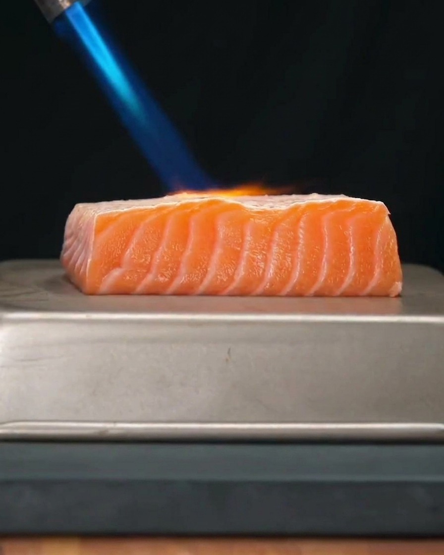 Un filete de salmón fresco siendo flameado con una llama azul de un soplete de cocina.