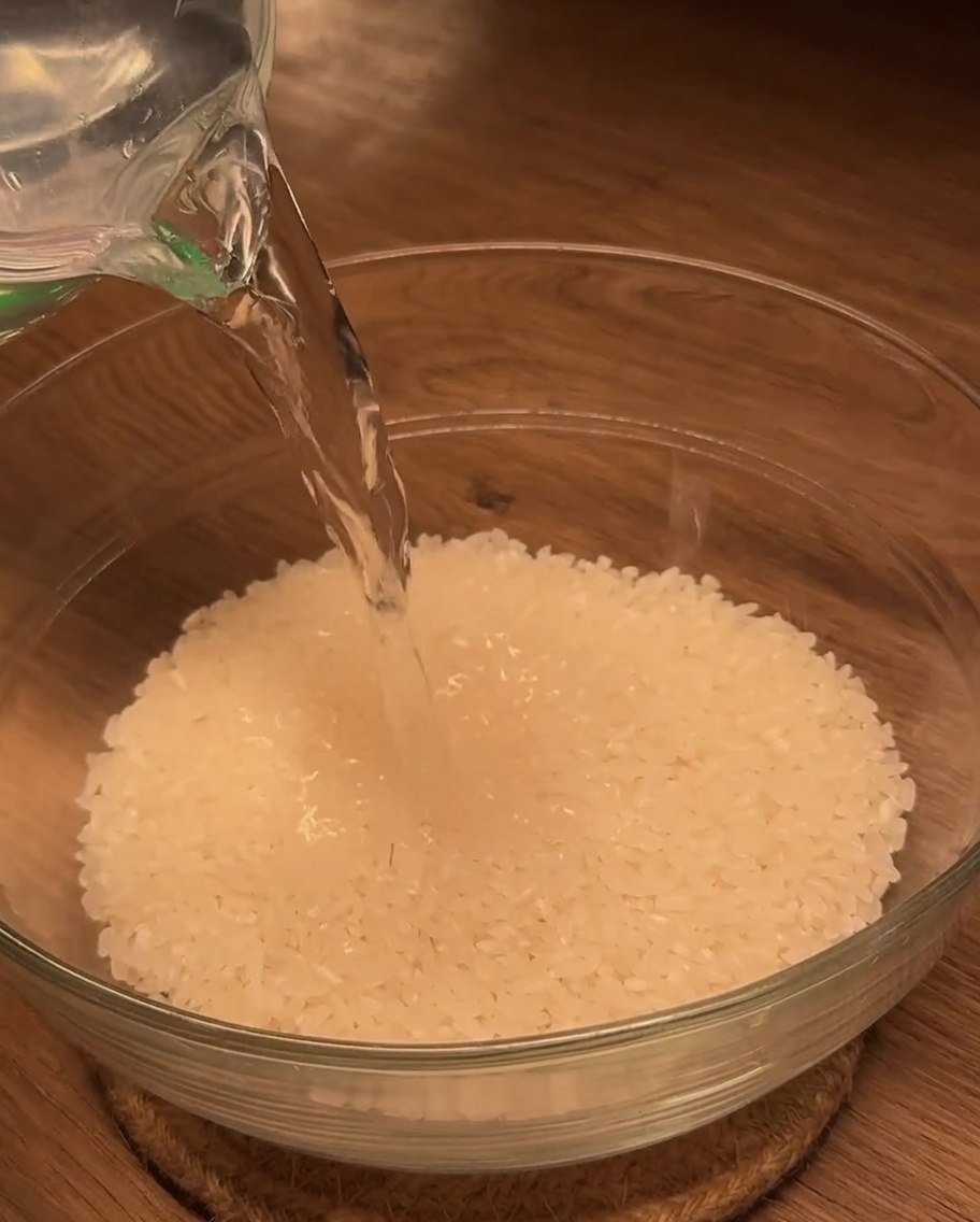 Agua vertida desde una taza medidora en un cuenco de cristal lleno de arroz blanco.