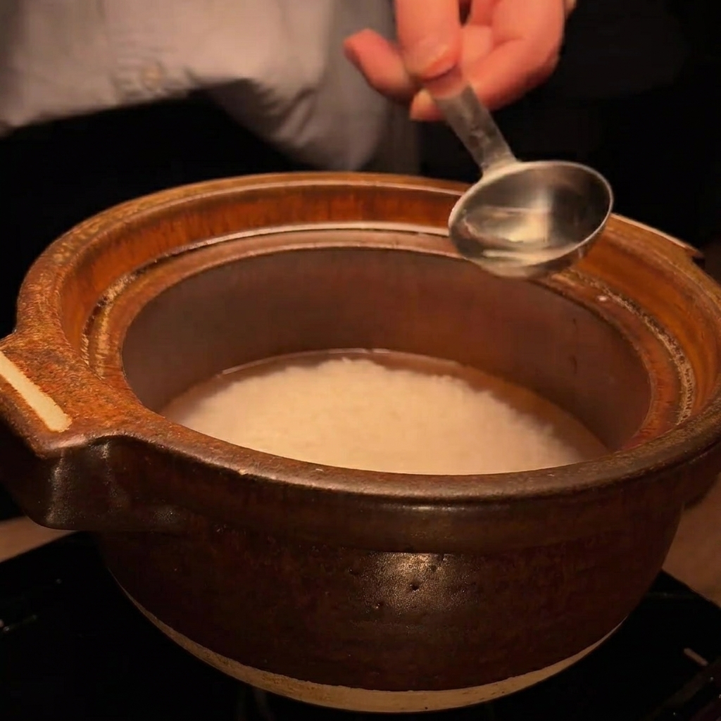 Una cuchara de metal vertiendo una pequeña cantidad de sake en la olla de barro con arroz.