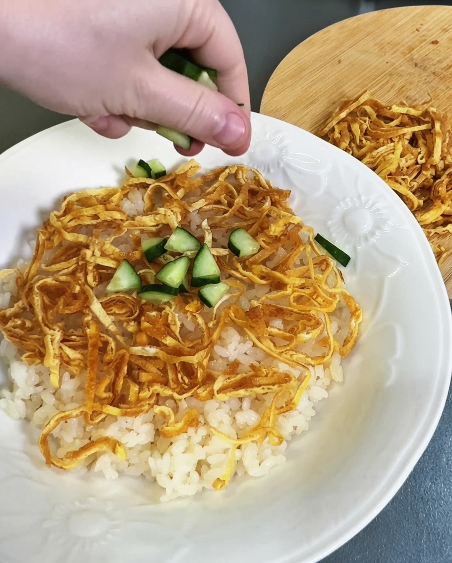 Pepino verde en cubitos siendo esparcido sobre la colorida capa de arroz avinagrado y tiras de huevo.