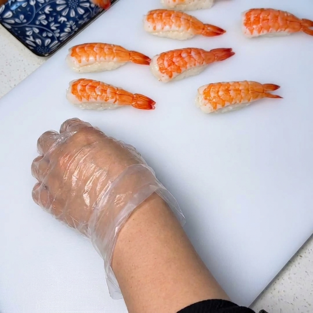 Primer plano de un langostino cocido colocándose sobre una bola de arroz, mientras unas manos con guantes presionan para darle forma a la pieza de sushi.