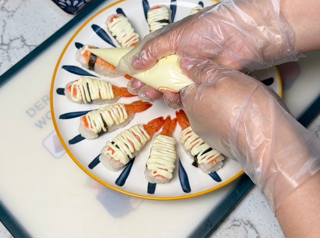 Primer plano de una manga pastelera distribuyendo mayonesa sobre las piezas de sushi de langostino dispuestas en un plato.