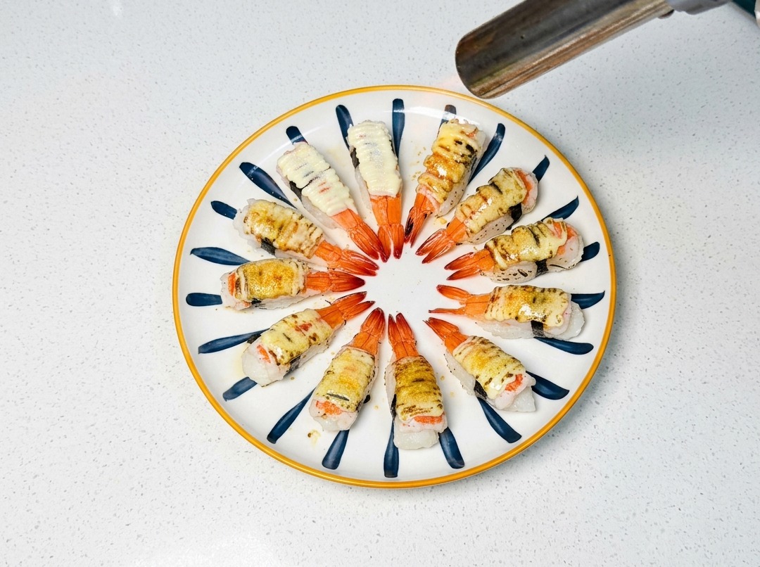 Un soplete de cocina dorando el azúcar y la cobertura de mayonesa sobre las piezas de sushi de langostino.