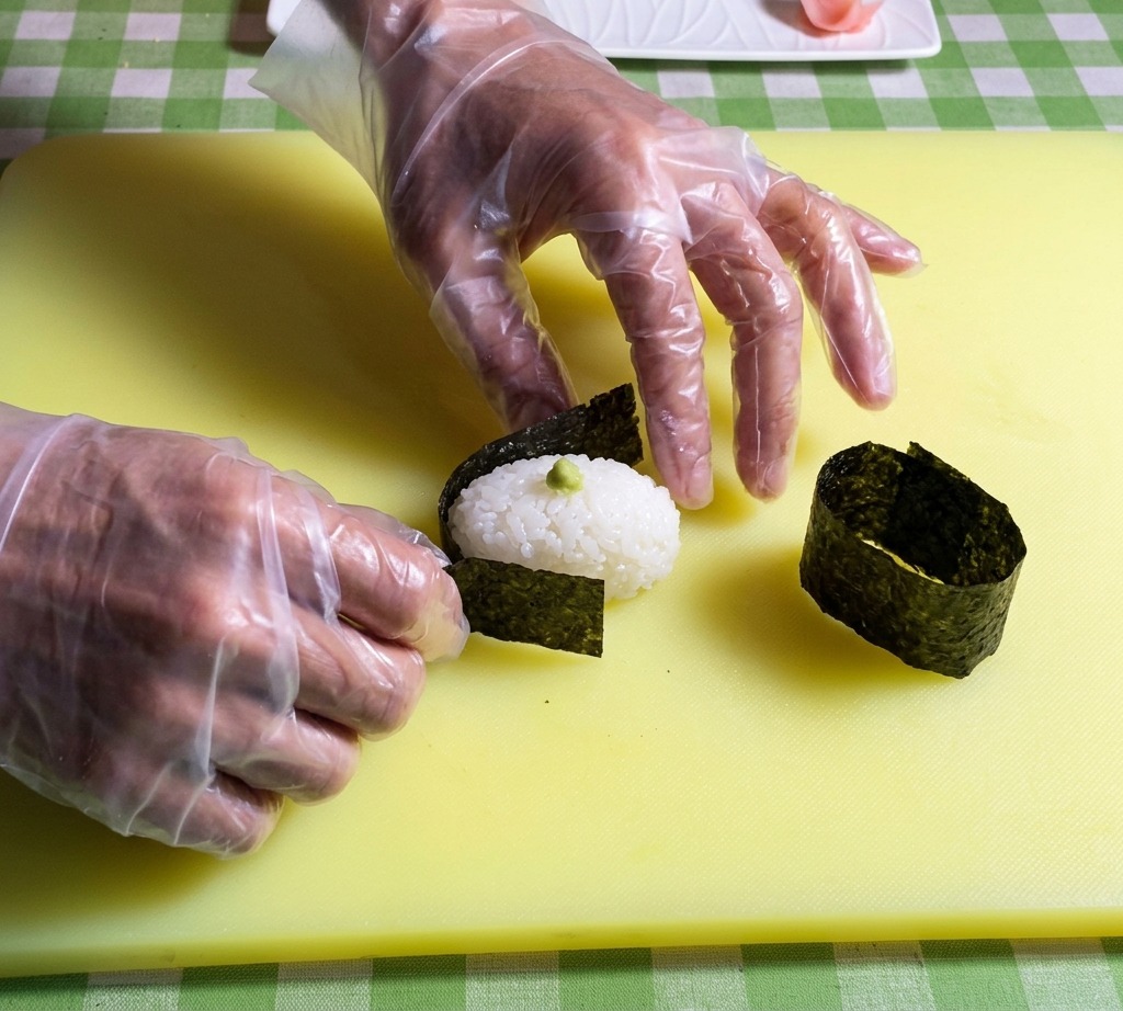 Manos colocando una tira de alga nori oscura alrededor del borde de una base de arroz ovalada.