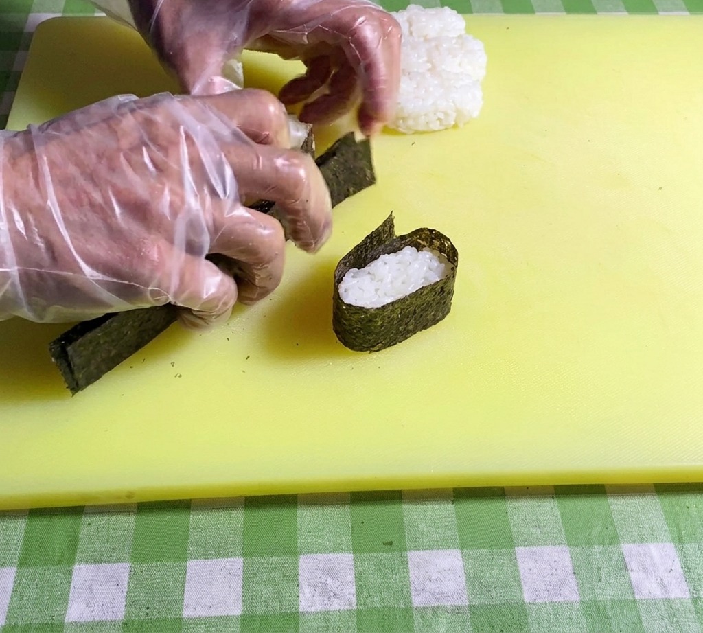 Primer plano de manos presionando los extremos de una tira de nori para asegurarla alrededor de la base de arroz.