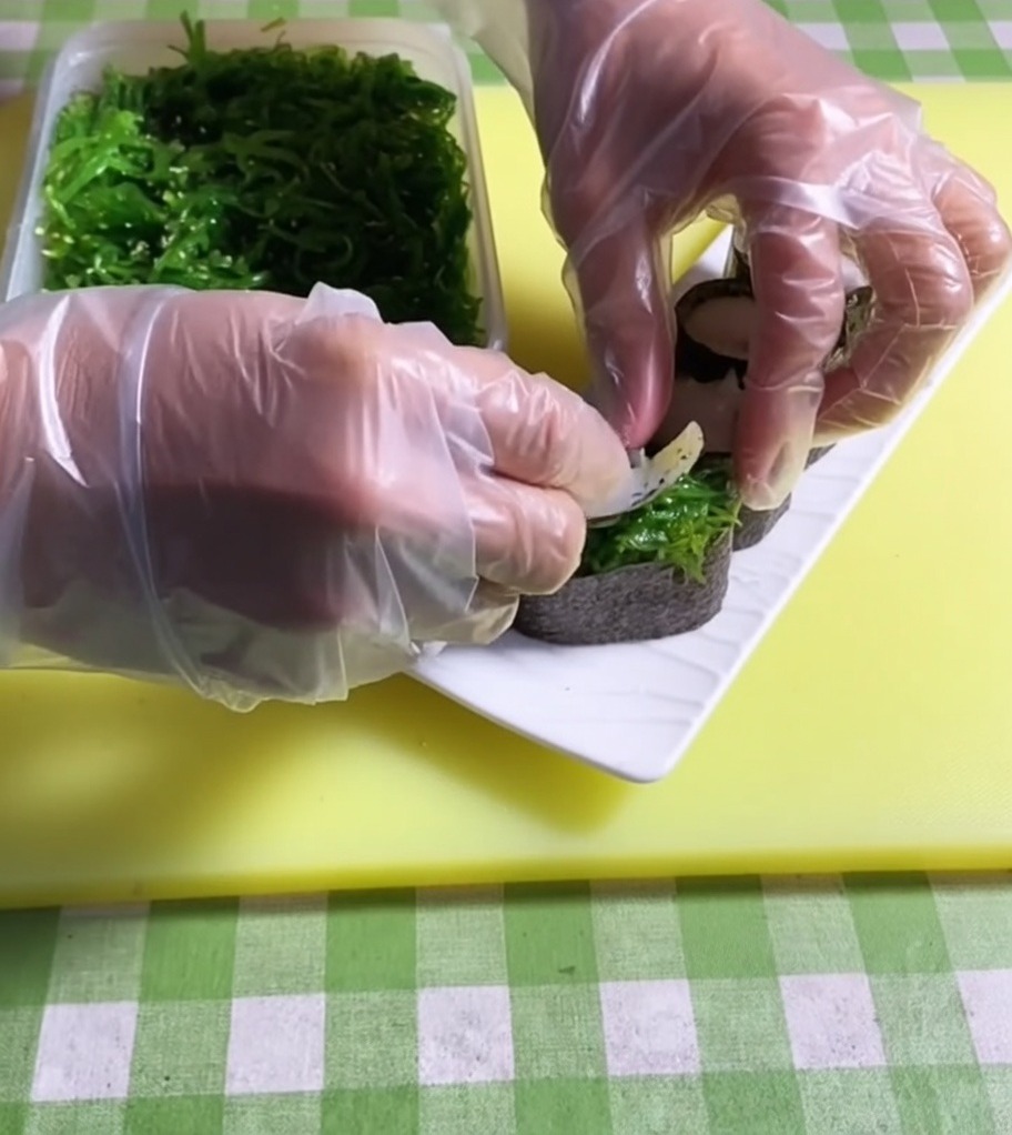 Pinzas colocando una porción verde vibrante de ensalada de algas sobre una base de sushi.