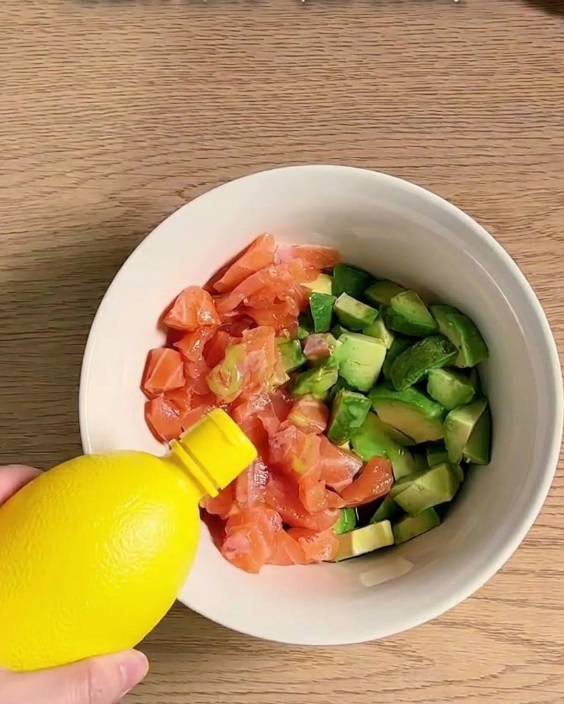 Un tazón lleno de trozos de salmón y aguacate siendo sazonado y mezclado con varios ingredientes.