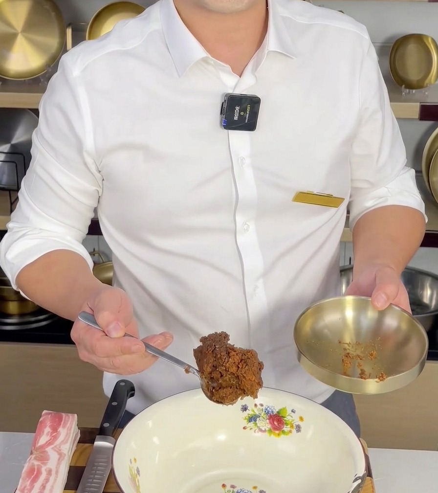 Una cucharada de Doenjang siendo agregada a un tazón de mezcla blanco, lista para combinarse con otros ingredientes.