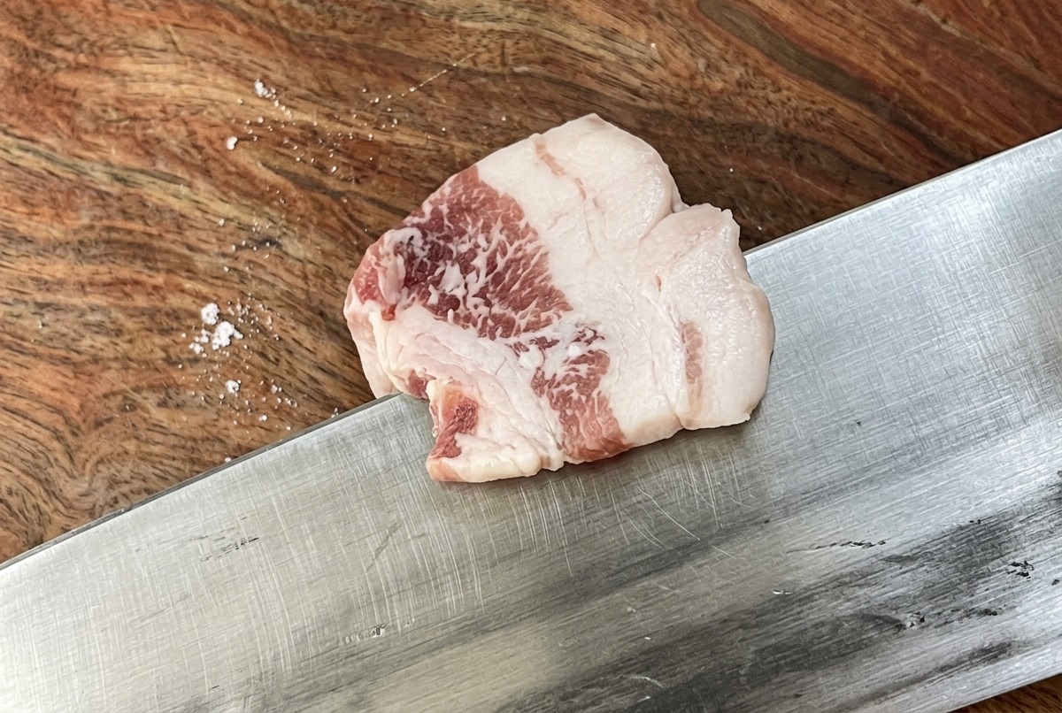 Un cuchillo afilado cortando una pieza de panceta cruda en tiras finas y uniformes sobre una tabla de cortar de madera.
