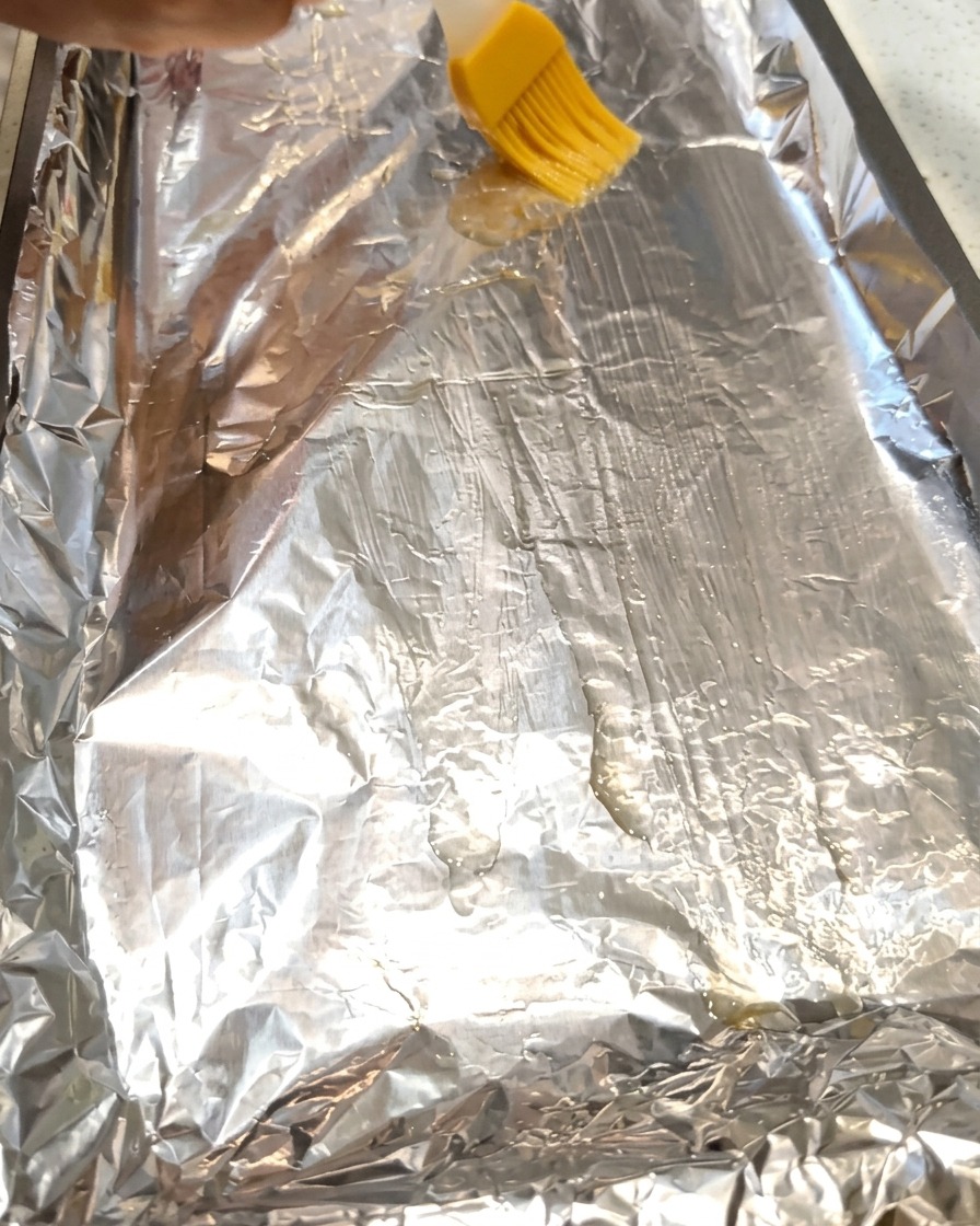 Un pincel esparciendo aceite de cocina sobre una hoja de papel aluminio en una bandeja de horno.