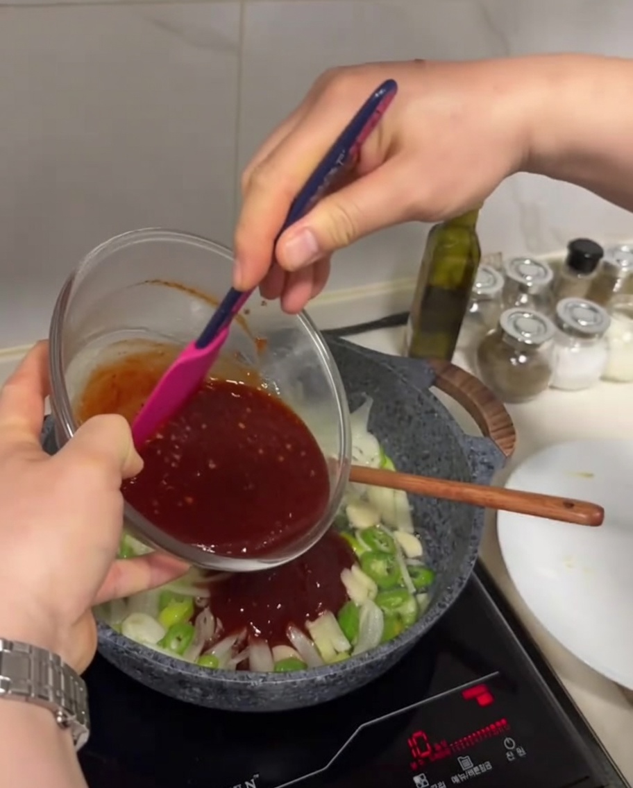 Salsa de color rojo oscuro siendo vertida desde un tazón a una sartén con verduras salteadas.